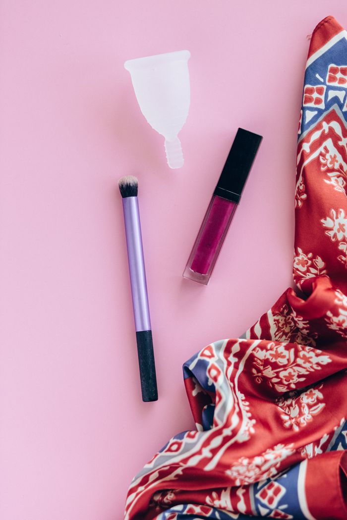 Creative composition of menstrual cup, lipstick, and makeup brush on pink background for beauty and wellness.